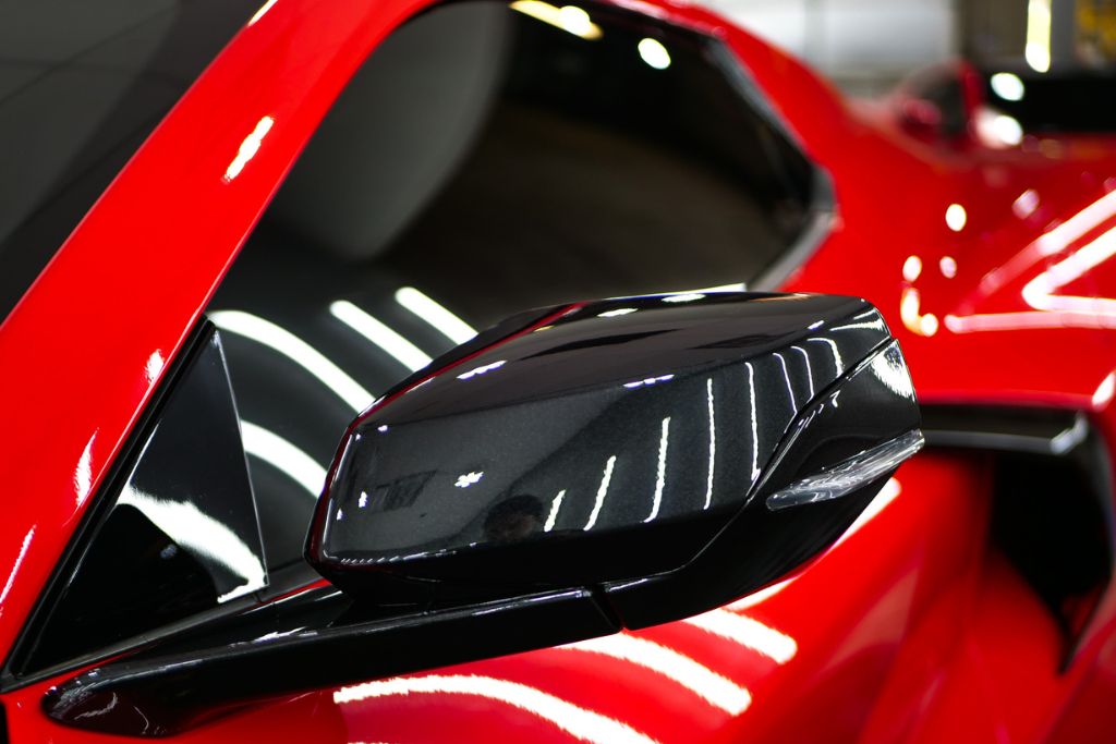 A close-up of a red sports car's side mirror and tinted window, reflecting bright LED lights. The deep black ceramic window tint enhances privacy and reduces glare, offering a sleek contrast against the glossy red paint. The reflection highlights the smooth finish of the tint application.