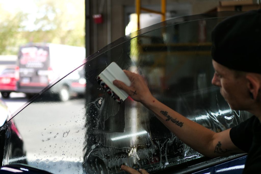 A professional installer applies ceramic window tint to a car door, using a squeegee to smooth out bubbles and ensure a perfect fit. The technician, wearing a black cap and showcasing tattooed arms, works in a well-lit garage with vehicles visible in the background. The process highlights the precision and quality of professional ceramic tinting services.