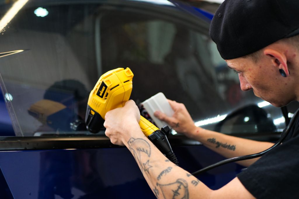 A professional window tint installer uses a heat gun and squeegee to apply ceramic tint to a car window. The technician, wearing a black cap and showcasing tattooed arms, carefully smooths the film to ensure a flawless, long-lasting application. This process highlights the superior durability and longevity of ceramic window tint, even in extreme heat conditions.