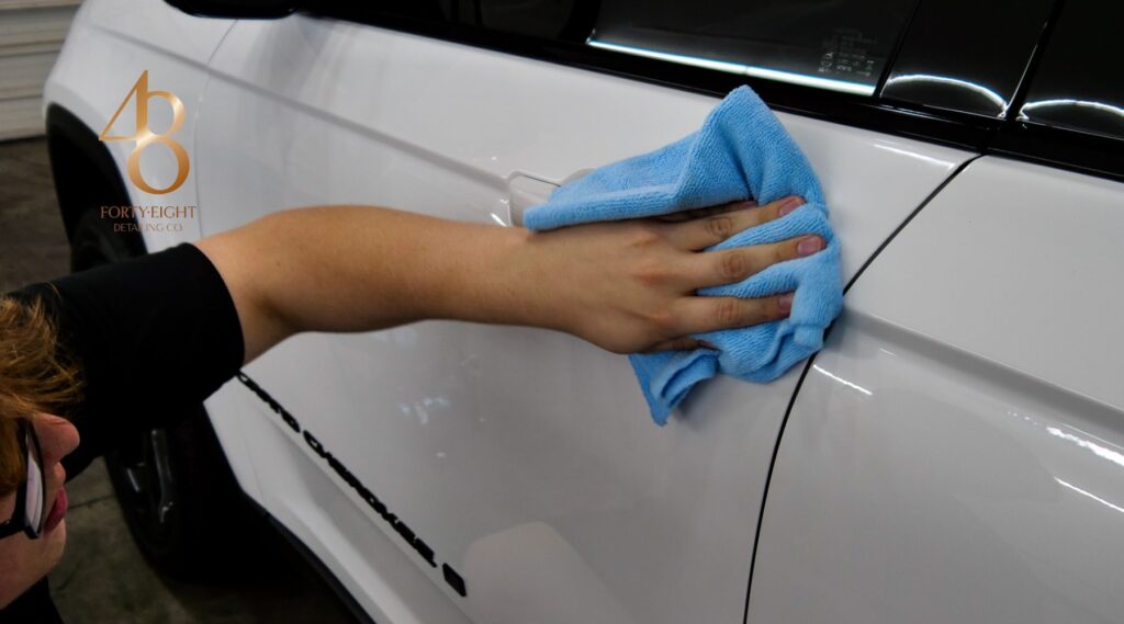 A detailer wipes down the glossy white exterior of a vehicle using a blue microfiber towel, ensuring a clean and polished finish. The "48 Detailing" logo is visible in the background, indicating a professional ceramic coating or detailing service.