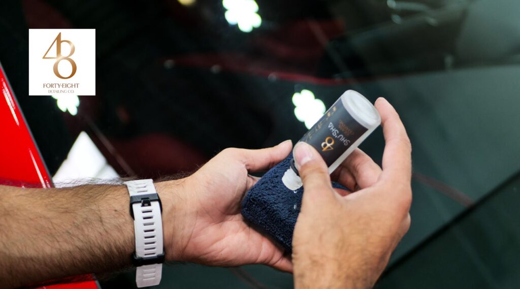 A person applies a ceramic coating product from Forty-Eight Detailing Co. onto a dark blue microfiber cloth. The bottle is labeled "SHUSHA" and is tilted as liquid is dispensed. The reflection of bright lights on a glossy car surface is visible in the background.