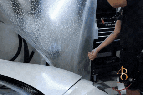 Detailing technician applying a large sheet of clear paint protection film over the hood of a white car, showcasing professional PPF installation included in 48 Detailing’s protection packages.