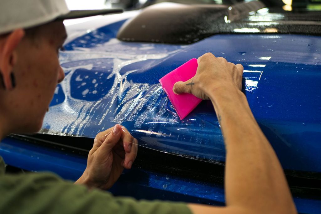 Technician applying paint protection film to the hood of a blue vehicle using a pink squeegee, ensuring smooth adhesion and long-term protection.