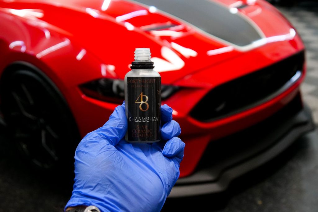 Gloved hand holding a bottle of 48 Detailing ceramic coating in front of a shiny red sports car, illustrating the high-gloss finish and protective benefits of ceramic coating against sun damage, dirt, and water spots.