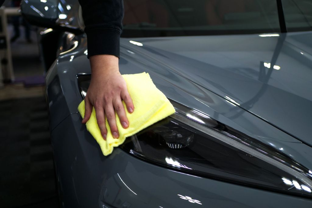 Hand using a bright yellow microfiber cloth to gently wipe the headlight area of a gray car, showing safe cleaning practices to protect car paint from bird droppings.