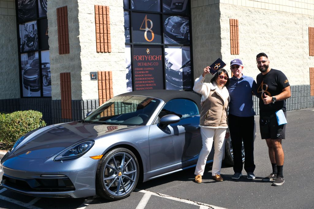 Happy couple posing with a detailing specialist beside their freshly coated silver Porsche convertible outside a detailing shop, representing the decision process when asking should I apply ceramic coating.