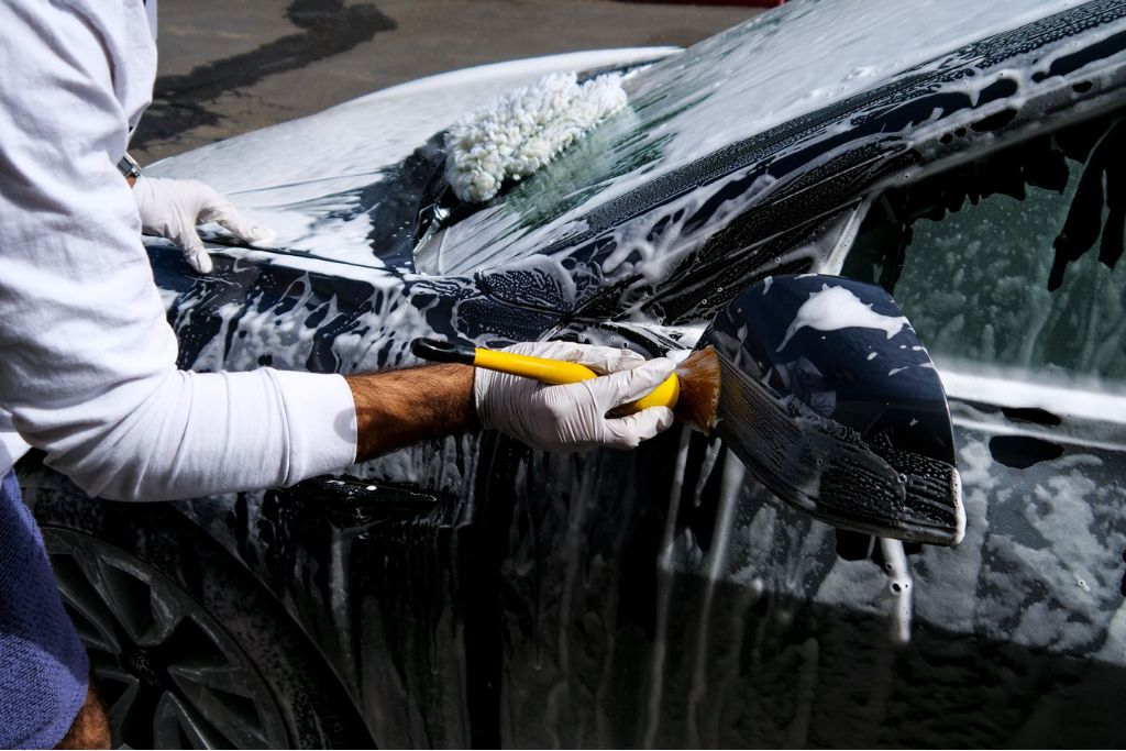 Person detailing a soapy black car with a soft brush around the side mirror, demonstrating preventative care to protect car paint from damage like bird droppings.