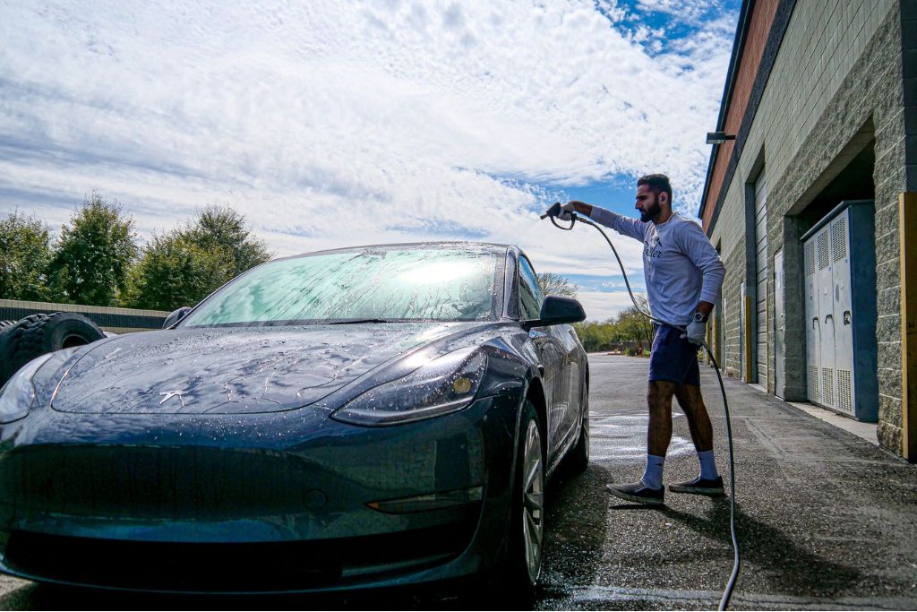 Man rinsing a dark gray Tesla outside a detailing shop, showing how maintaining clean windows helps preserve UV protection in car window tints.
