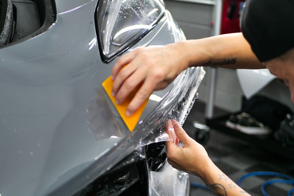 A professional installer applies Paint Protection Film (PPF) to a car’s front bumper, using a squeegee to smooth the transparent film over the surface. This image visually explains How PPF Works by showing the detailed film installation process that protects the vehicle’s paint.