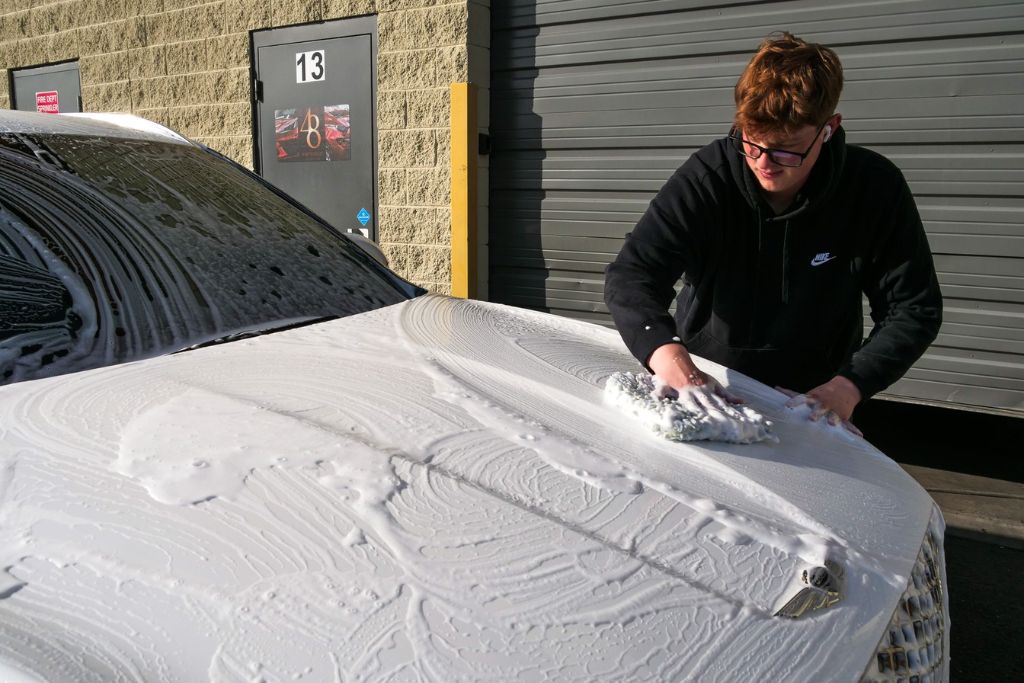 Person hand-washing a foam-covered white luxury car with a soft mitt, demonstrating proper maintenance that helps determine how long ceramic coating lasts.