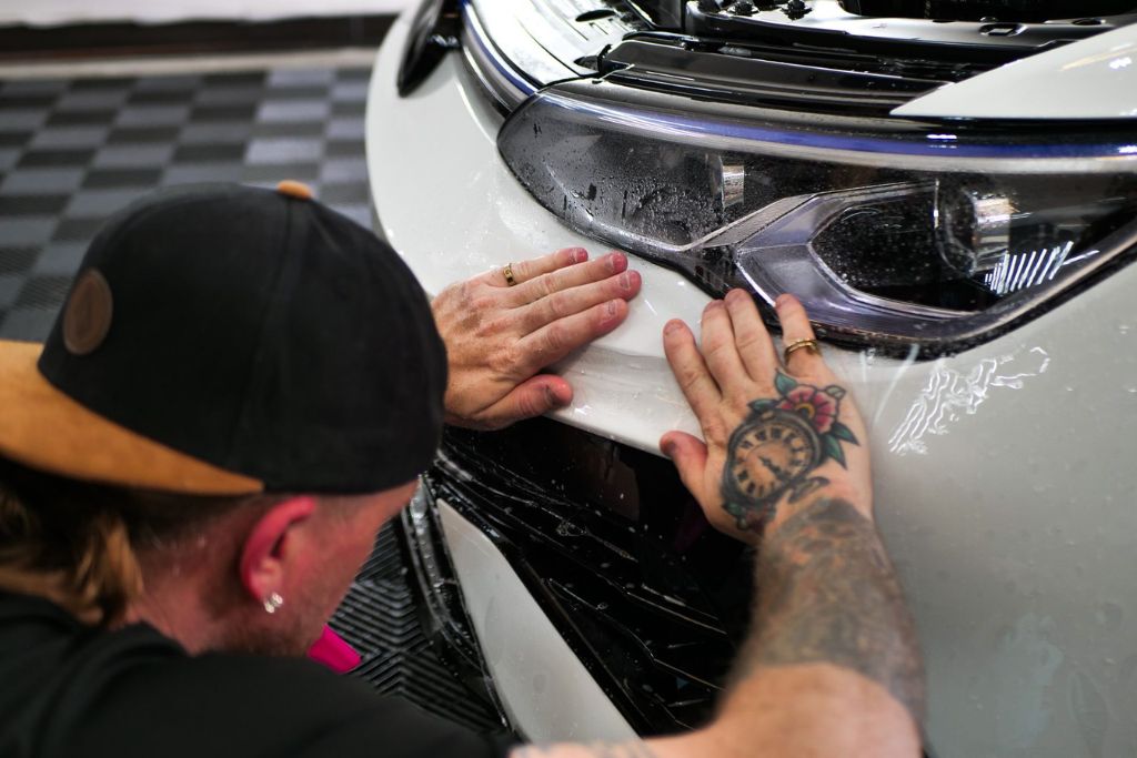 Technician carefully applying paint protection film to the front bumper of a white car, focusing on high-impact areas vulnerable to rock chips.