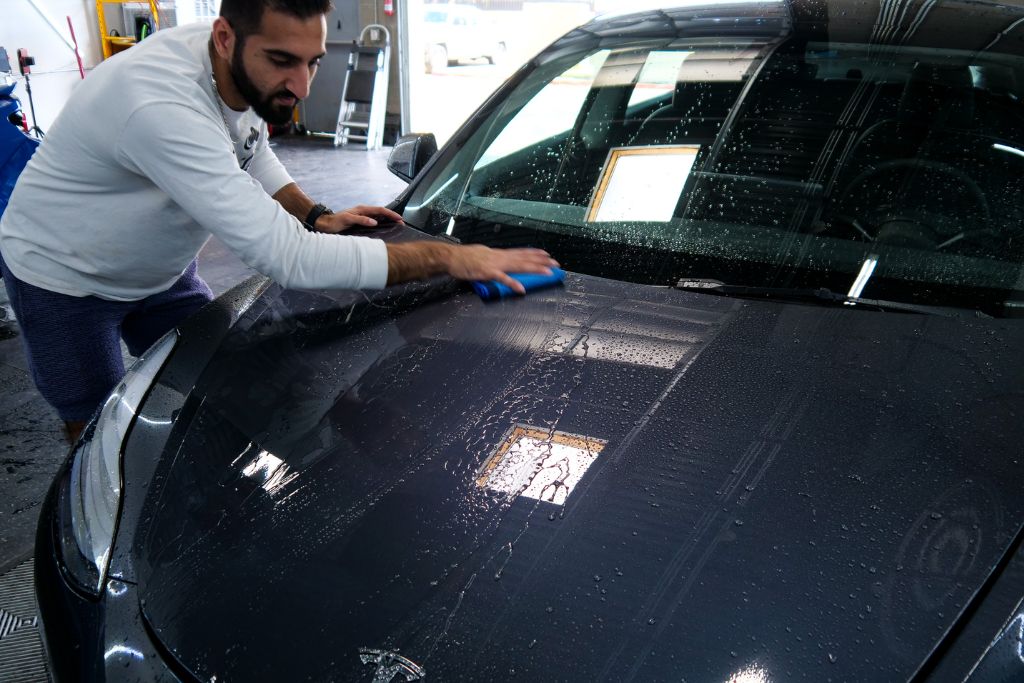 Detailer using a blue applicator pad to inspect and clean the wet hood of a dark-colored car, checking for hydrophobic performance and signs of ceramic coating wear.