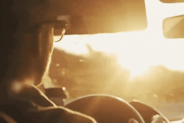 Driver wearing sunglasses steers a car toward a bright sunset, with sunlight streaming through the windshield and creating a warm golden glow inside the cabin.