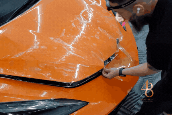 Technician applying transparent bra paint protection film to the hood of an orange sports car, ensuring precise fit and long-term scratch prevention.