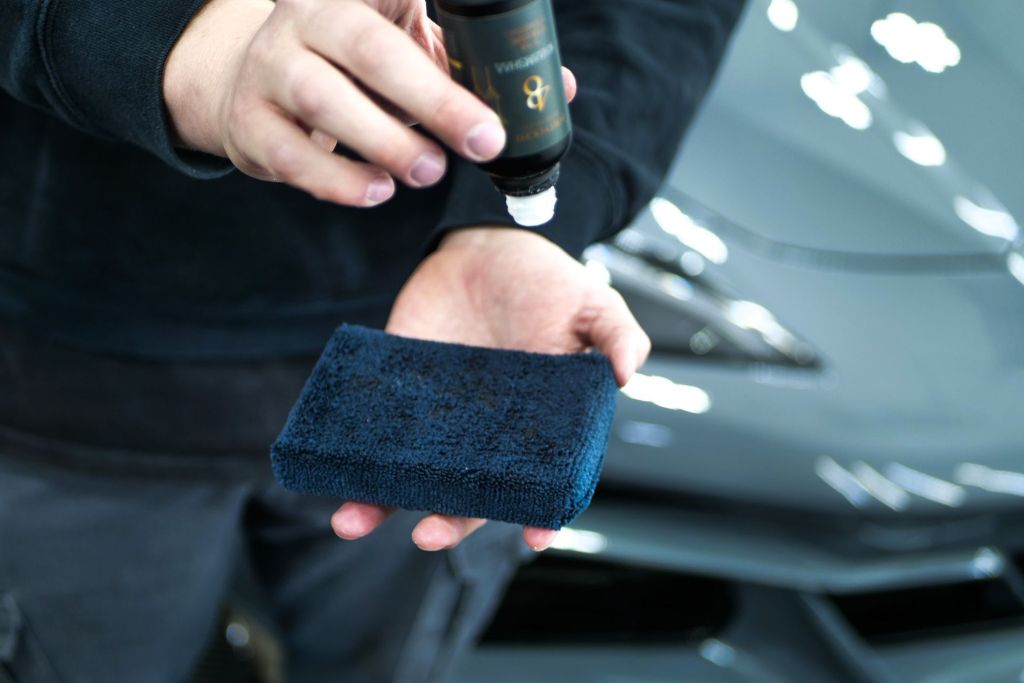 Person applying ceramic coating onto a blue microfiber applicator pad in front of a sleek gray car, illustrating the process that affects how long ceramic coating lasts.