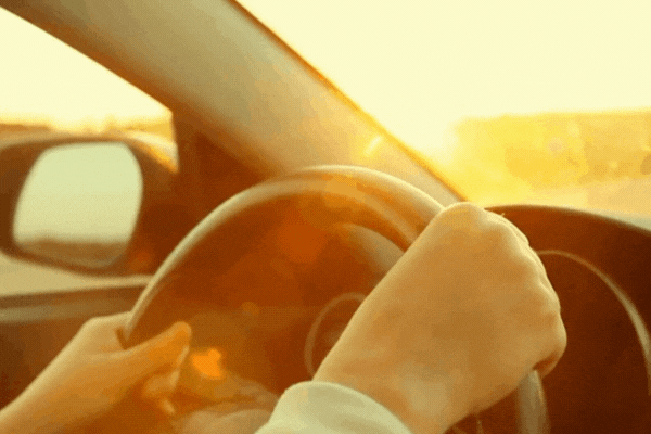 Close-up of hands gripping a steering wheel while driving into bright sunlight, representing the need for UV protection for both driver and car interior.
