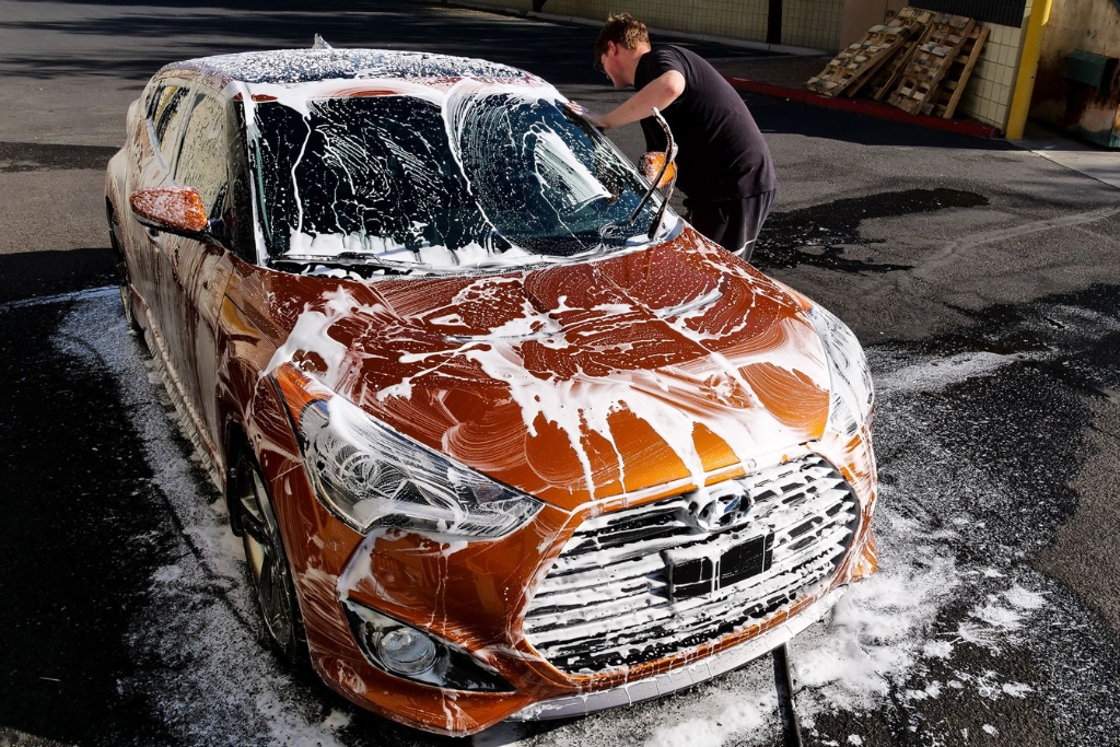 A man hand-washing a foam-covered orange car in bright Arizona sunlight, illustrating How to Prevent Water Spots through proper drying and detailing techniques even with a ceramic coating