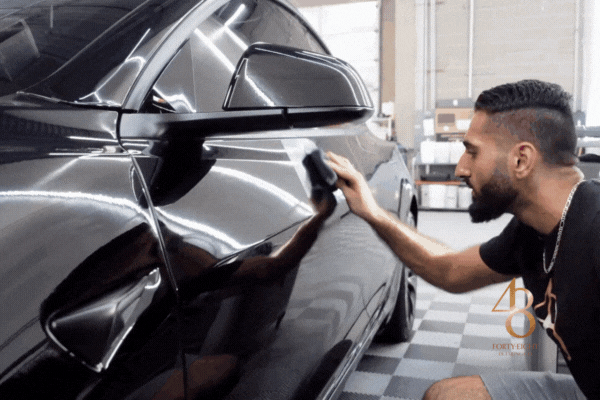 A detailing technician from 48 Detailing carefully applies ceramic coating to the side of a sleek black car inside a professional shop, enhancing the paint’s depth and shine while adding long-lasting protection.
