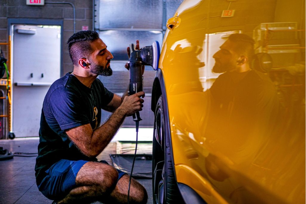 Detailing technician uses a dual-action polisher on a bright yellow car inside a garage, creating a glossy reflection on the vehicle’s surface—illustrating a common step when combining Paint Protection Film and Ceramic Coating for full-body protection and shine.