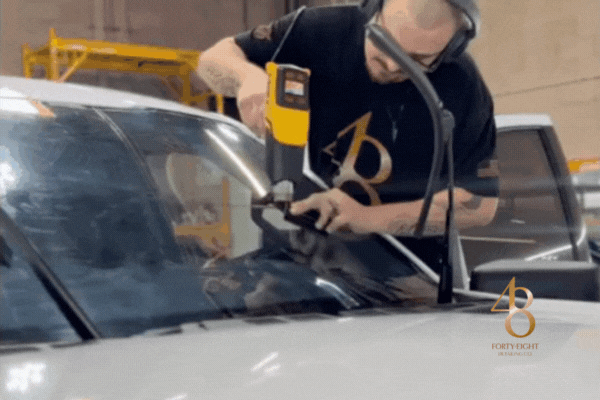 An automotive tint technician at 48 Detailing in Surprise Arizona uses a heat gun to apply window film to a vehicle’s windshield with precision. The scene showcases the professional skill and clean environment that defines why drivers choose to work with 48 Detailing for expert-level ceramic tint and vehicle protection services.
