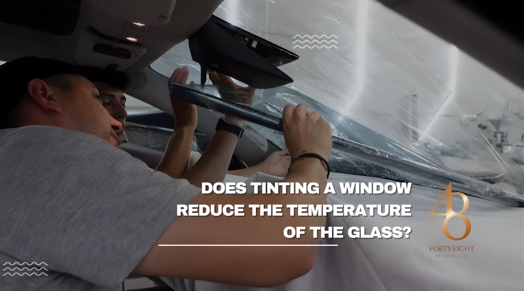 Two men apply a sheet of window tint film to the inside of a car windshield, demonstrating the tinting process. Text overlay reads, "Does tinting a window reduce the temperature of the glass?" with the Forty-Eight Detailing Co. logo in the bottom right.