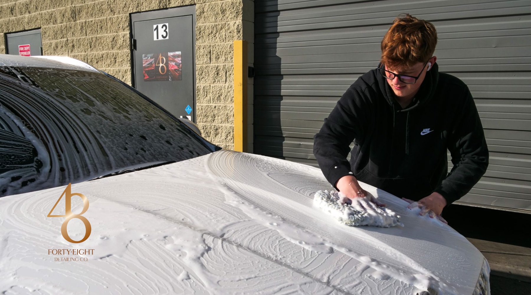 A man washing a white car covered in thick foam outside a detailing shop in Surprise, AZ, demonstrating how to maintain ceramic coating in winter. The image features the 48 Detailing logo on the hood, emphasizing professional seasonal car care.