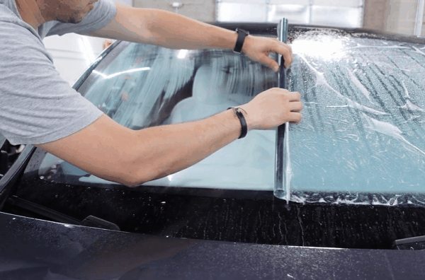 A technician applies a large sheet of window tint to a car windshield, smoothing it down with precision to block heat-causing solar radiation. Water and air bubbles are visible beneath the film during the installation process.