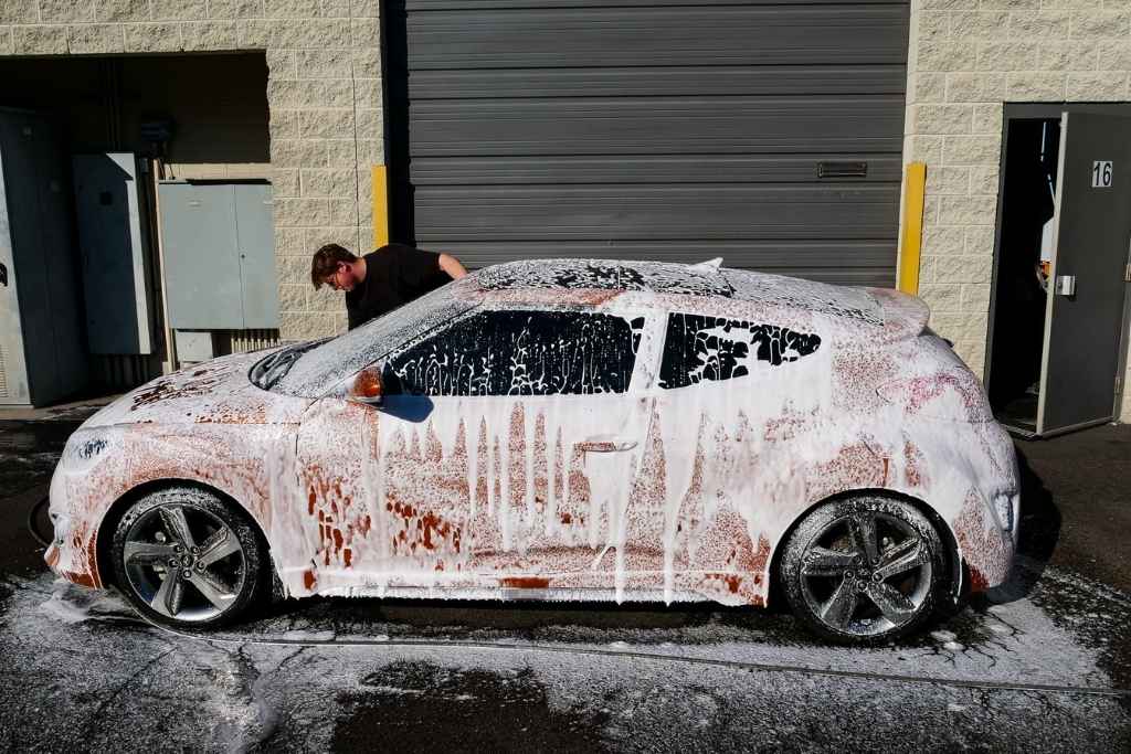 A red car fully covered in thick foam during a professional wash at an auto detailing shop in Surprise, AZ, with a technician working on the vehicle. This image highlights the expert winter ceramic coating maintenance offered by 48 Detailing.