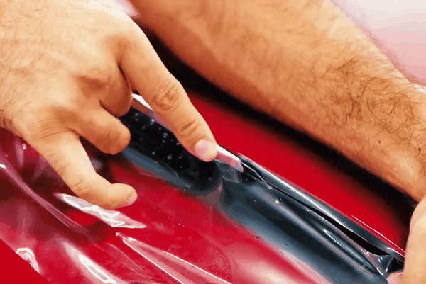 Technician pulling and positioning paint protection film over the hood and headlight of a red vehicle.