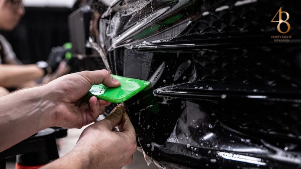 Technician using a green squeegee to apply paint protection film on the front bumper of a black car with a textured grille.