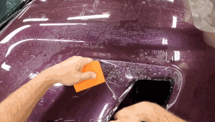 Hands applying and smoothing paint protection film on a wet purple car hood using an orange squeegee tool.
