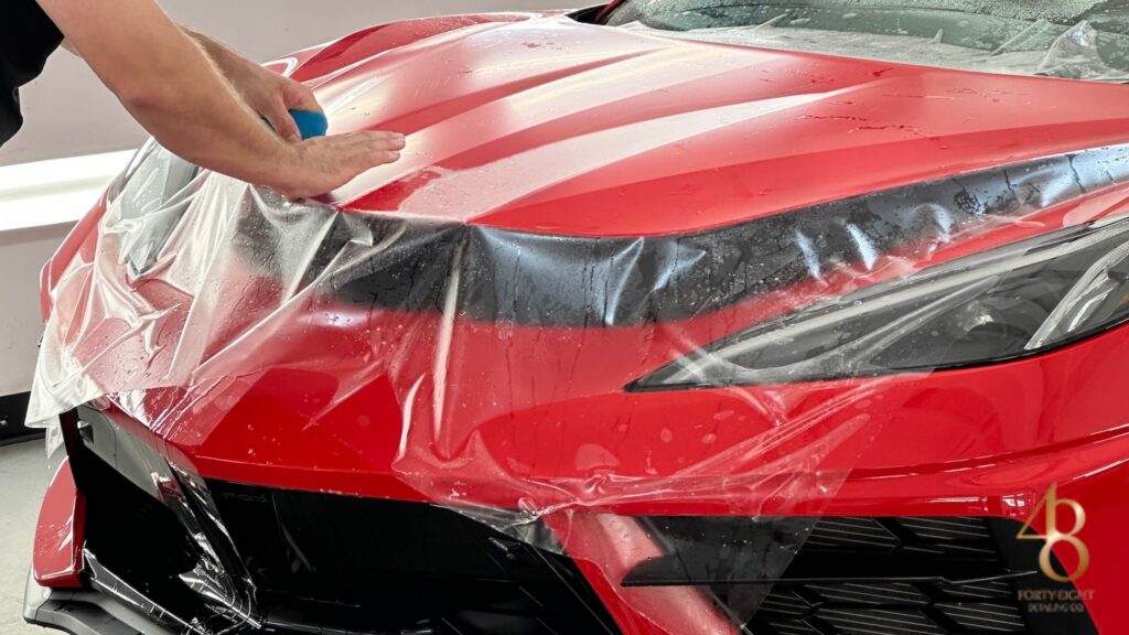 Installer smoothing transparent PPF across the hood of a red sports car using a blue squeegee. The paint protection film is laid over the headlights and front end to guard against road debris and surface damage. Water droplets beneath the film show the wet application process used for seamless PPF installation.