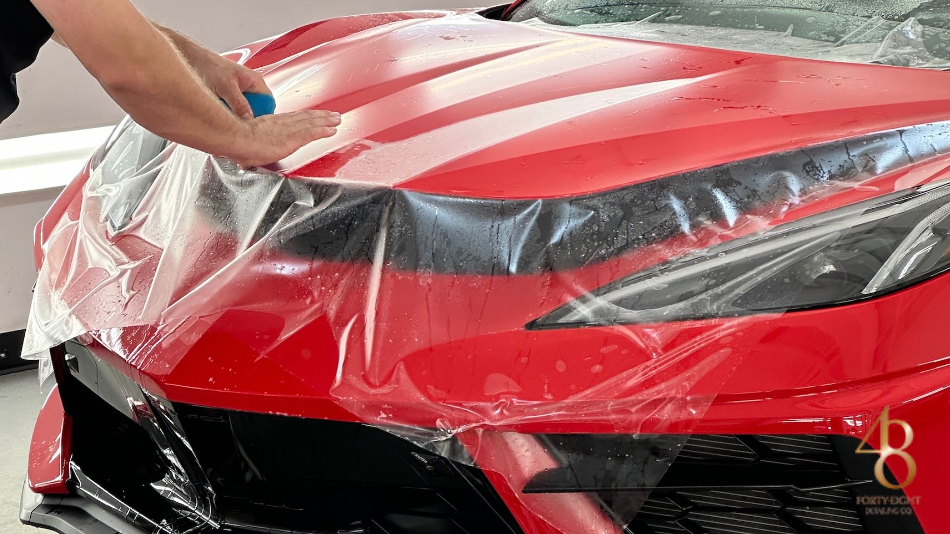 Installer smoothing transparent PPF across the hood of a red sports car using a blue squeegee. The paint protection film is laid over the headlights and front end to guard against road debris and surface damage. Water droplets beneath the film show the wet application process used for seamless PPF installation.