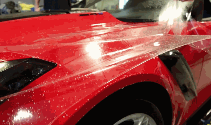 Clear paint protection film being carefully laid across the hood of a red car covered in water droplets during installation. The transparent paint protection film is stretched smoothly over the glossy surface to shield the finish from chips and scratches.