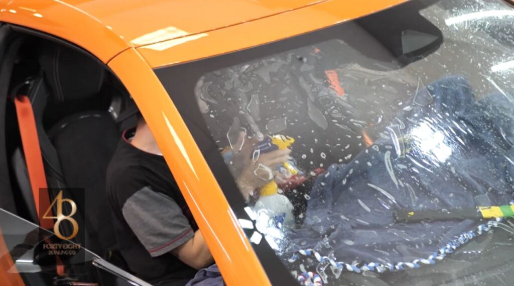 Technician installing window tint film on the windshield of an orange sports car at 48 Detailing.