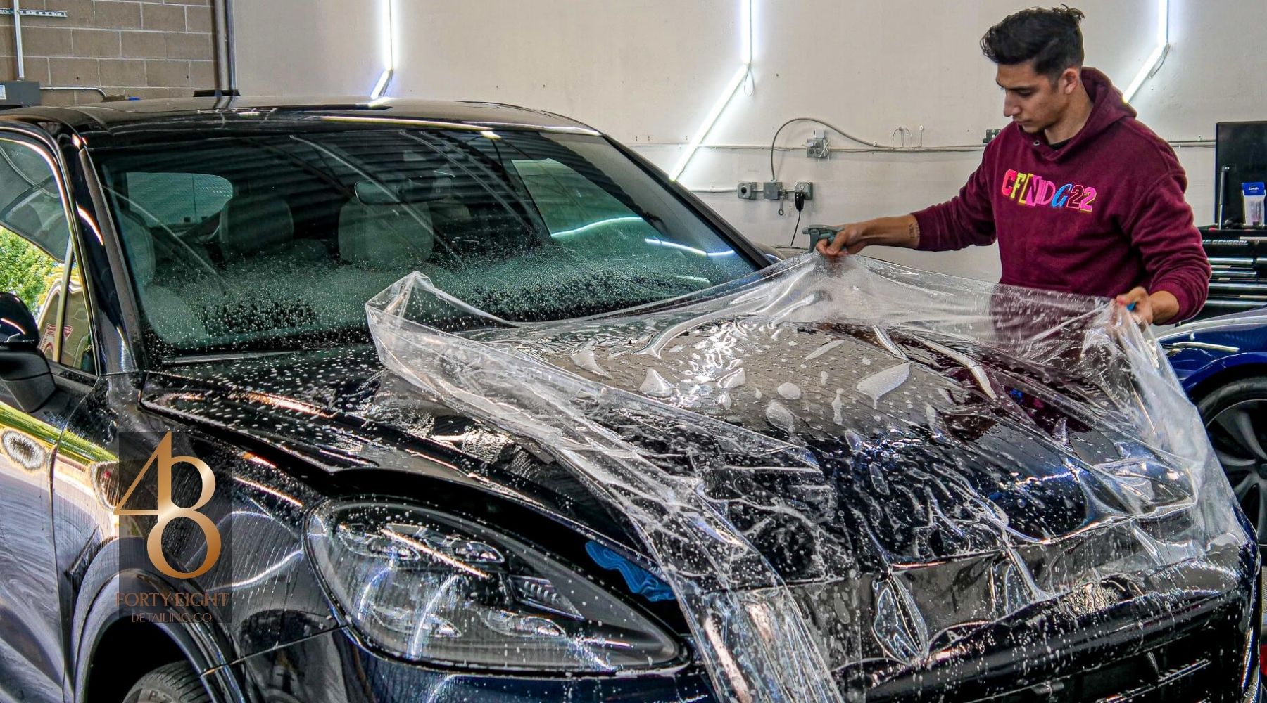 48 Detailing technician installing clear paint protection film on the hood of a black vehicle in a professional detailing studio.
