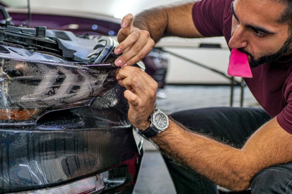 48 Detailing technician carefully trimming paint protection film around a vehicle’s headlight and front bumper.