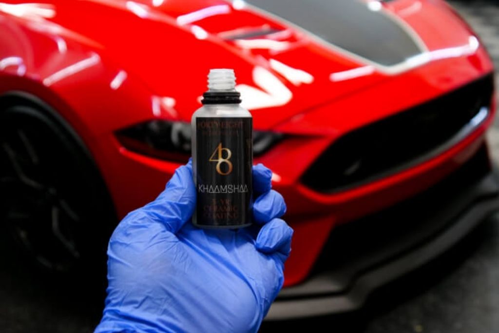 Technician holding a ceramic coating bottle in front of a glossy red Mustang at 48 Detailing.