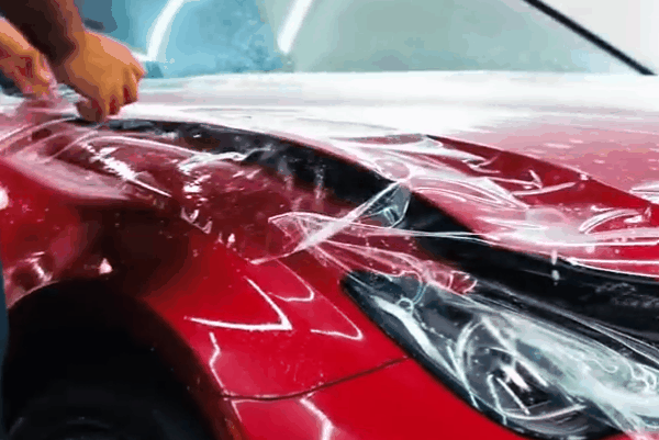 Technician applying clear paint protection film around the headlight and front fender of a red vehicle.