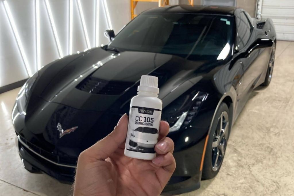 Hand holding a bottle of CC 105 ceramic coating in front of a black sports car inside a detailing studio.